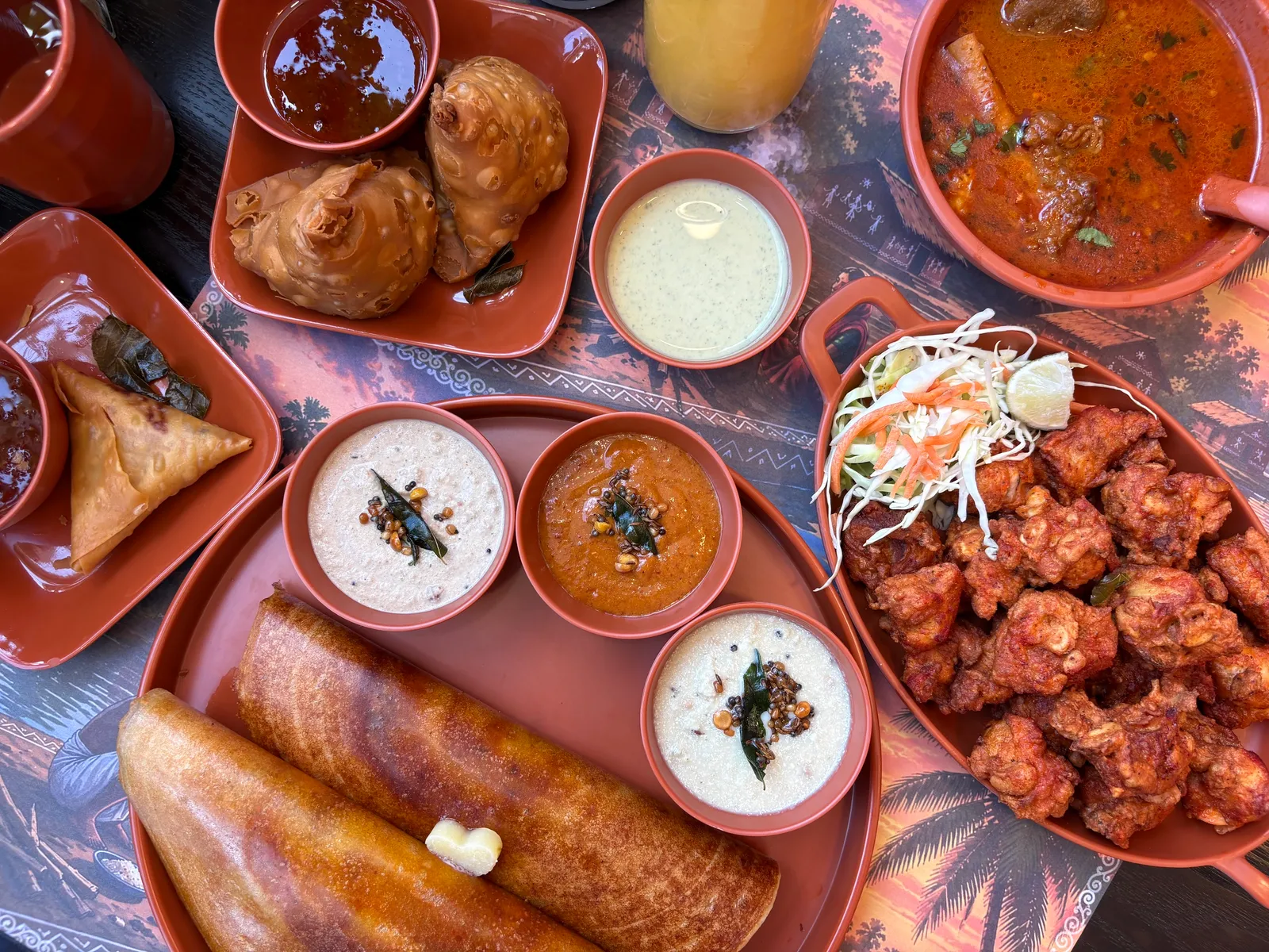 An overhead spread of dosas, samosas, fried chicken, curry and chutneys across a laid table