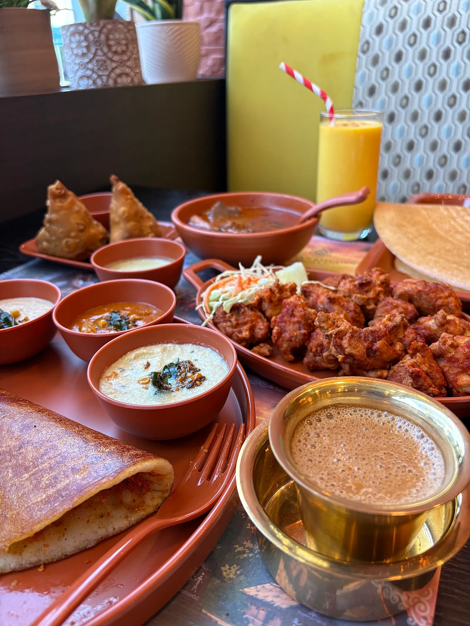 A table set with folded dosa, samosas, chutneys, mango lassi and filter coffee in a steel tumbler