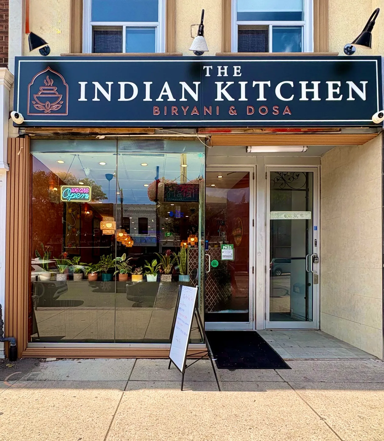 The Indian Kitchen storefront on Yonge Street, navy Biryani and Dosa sign above a plant-filled window with a neon open sign